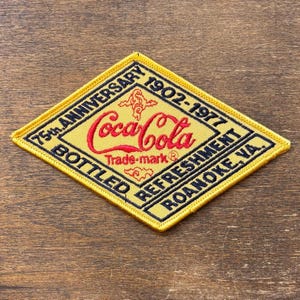 May include: A yellow and black embroidered patch in a diamond shape. The patch features the Coca-Cola logo in red, with text indicating the 75th anniversary, the years 1902-1977, and the location Roanoke, VA.