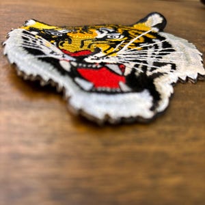 May include: An embroidered tiger head patch featuring vibrant yellow, black, and white thread detailing. The design depicts a tiger with an open mouth, showing red and white accents, long white whiskers, and sharp teeth, finished with a white border.
