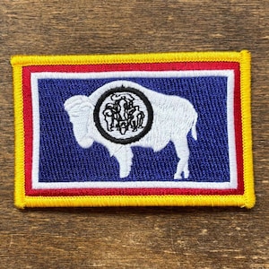 Puede incluir: Parche bordado con el diseño de la bandera del estado de Wyoming. El parche tiene un fondo azul con un bisonte blanco en el centro, rodeado por un emblema negro. El parche es rectangular con un borde amarillo, bordes interiores rojos y blancos.