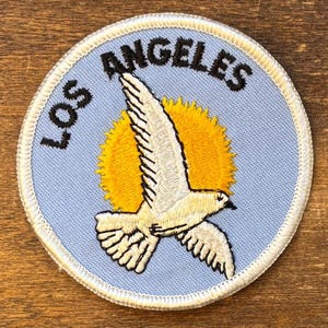 May include: Circular embroidered patch with a white seagull flying in front of a yellow sun on a light blue background. The words "LOS ANGELES" are arched above the bird in black. The patch has a white border.