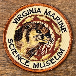May include: Embroidered patch featuring a detailed otter illustration. The patch has a circular design with the text "VIRGINIA MARINE SCIENCE MUSEUM" in black lettering on a cream background, framed by a brown border.