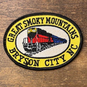 Op de afbeelding: Een ovale geborduurde patch met een rode, zwarte en blauwe treinlocomotief op een witte achtergrond. De gele rand bevat de zwarte tekst GREAT SMOKY MOUNTAINS en BRYSON CITY NC. De patch heeft een zwart gestikte randafwerking.