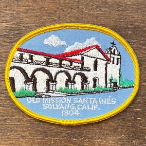 May include: Embroidered patch featuring a depiction of Old Mission Santa Inés in Solvang, California. The patch has a blue background with a detailed illustration of the mission building, red roof, and text that reads "Old Mission Santa Inés Solvang, Calif. 1804."