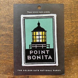 Pode incluir: Um patch de Point Bonita com um farol preto e amarelo sobre um fundo verde-azulado, com as palavras "POINT BONITA" em branco. O patch está em um cartão preto com o texto "THE GOLDEN GATE NATIONAL PARKS".