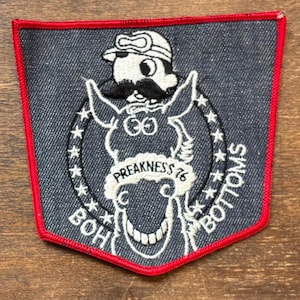 May include: Embroidered patch with a denim background and a red border. The patch features a cartoon donkey wearing goggles and a mustache, with the text "PREAKNESS 76" and "BOH BOTTOMS" in white.