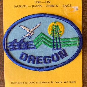 Puede incluir: Un parche bordado azul y blanco con una gaviota volando sobre una cordillera verde con un sol amarillo y la palabra "OREGON" en letras azules. El texto "USE - ON JACKETS - JEANS - SHIRTS - BAGS" está impreso en el fondo amarillo.