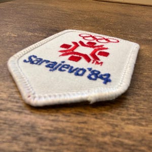 Puede incluir: Un parche blanco con un dise&ntilde;o rojo y azul. El parche presenta los anillos ol&iacute;mpicos y el texto "Sarajevo '84" en azul. El dise&ntilde;o incluye una figura estilizada y el texto "TM". El parche tiene un borde blanco.