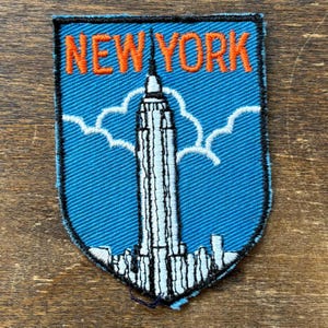 Peut inclure: Écusson brodé représentant l'horizon de New York, avec l'Empire State Building. L'écusson a un fond bleu, des nuages blancs et les mots "NEW YORK" en orange. L'écusson est rectangulaire avec des bords arrondis.
