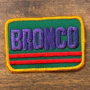 May include: Embroidered patch with the word "BRONCO" in purple, stitched onto a green background. Two red horizontal stripes are below the text. The patch has a yellow border.