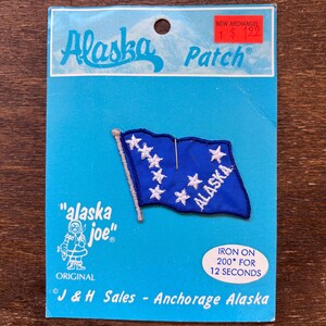 以下が含まれることがあります： アラスカの旗をデザインした青と白のアイロン接着パッチ。パッチは白い星模様と白い文字で「ALASKA」と書かれています。パッチは「Alaska Patch」と「J & H Sales - Anchorage Alaska」の文字が書かれた青いカードにパッケージされています。