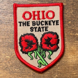 Pode incluir: Patch bordado com o estado de Ohio. O patch tem um fundo bege com uma borda vermelha. A palavra "OHIO" está em letras vermelhas em bloco acima do texto "THE BUCKEYE STATE". Duas flores vermelhas com centros pretos e caules verdes estão na parte inferior.
