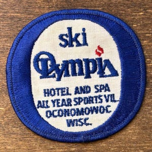 Könnte beinhalten: Ein kreisförmiger, gestickter Aufnäher mit blauem Rand und weißer Mitte. Der Text "ski Olympia" ist in Blau gehalten, wobei "Olympia" in einer stilisierten Schriftart geschrieben ist. Zusätzlicher Text lautet "HOTEL AND SPA ALL YEAR SPORTS VIL OCONOMOWOC WISC."