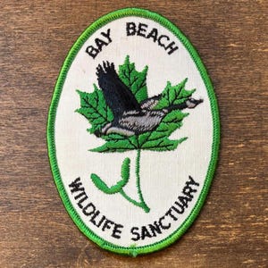 Op de afbeelding: Ovale patch met een groene rand en de woorden "BAY BEACH WILDLIFE SANCTUARY". De patch toont een groen esdoornblad met een zwarte en grijze vogel in vlucht.