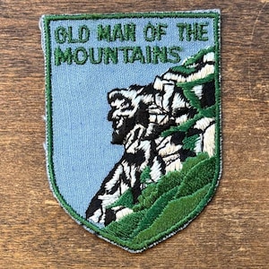 May include: Embroidered patch featuring the text "OLD MAN OF THE MOUNTAINS" above an illustration of a mountain with a face. The patch has a light blue background, green border, and green and black mountain details.