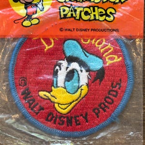 Puede incluir: Un parche bordado rojo y azul con Donald Duck y el texto "Walt Disney Prods." y "Donald" alrededor del borde.
