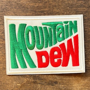Op de afbeelding: Geborduurde patch met de woorden "Mountain Dew" in groen en rood. De tekst staat op een witte rechthoekige achtergrond met een witte rand. Geschikt voor kleding of accessoires.