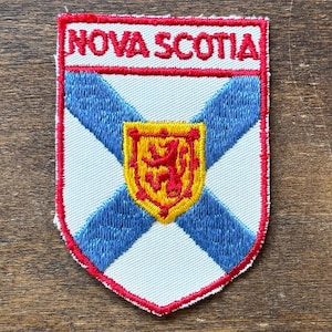 Könnte beinhalten: Ein gestickter Patch in Rot, Weiß und Blau mit der Aufschrift "NOVA SCOTIA" in roten Buchstaben. Der Patch zeigt das Wappen von Nova Scotia, das einen roten Löwen auf einem gelben Schild enthält.