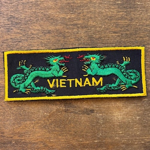 May include: Embroidered patch featuring two green dragons facing each other on a black background. The word "VIETNAM" is embroidered in yellow. The patch has a yellow border.