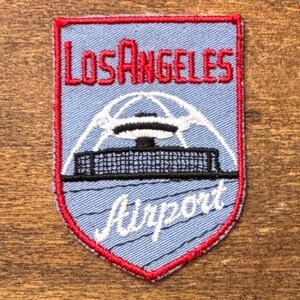 Peut inclure: Un écusson brodé en forme de bouclier avec un fond bleu clair et une bordure rouge. Il présente le texte rouge LOS ANGELES au-dessus d'un graphique blanc du Theme Building de LAX, avec le texte cursif blanc Airport sous la structure.
