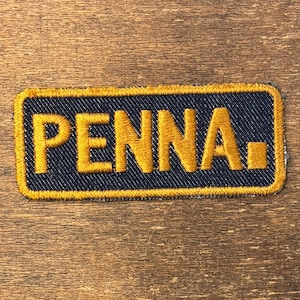 Pode incluir: Um emblema retangular bordado com um fundo azul escuro semelhante a ganga e uma borda dourada espessa. A palavra PENNA está cosida em letras maiúsculas douradas, seguida por um pequeno símbolo quadrado dourado no final do texto.