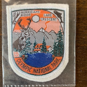 Op de afbeelding: Een rechthoekige patch met een landschapsontwerp, met de tekst "Hurricane Ridge" en "Lake Crescent". De patch bevat een afbeelding van een eland, bergen, een meer en de woorden "Olympic National Park". De patch zit in een doorzichtige plastic hoes.