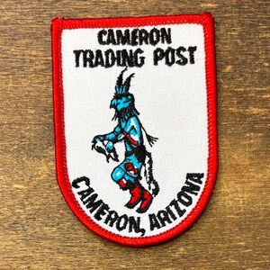 Może przedstawiać: Naszywka w kształcie tarczy z białym tłem i czerwoną obwódką. Zawiera czarny napis CAMERON TRADING POST oraz CAMERON, ARIZONA. W środku znajduje się wyhaftowana niebiesko-czarno-czerwona postać tańczącej osoby.