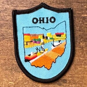 Può includere: Una toppa ricamata a forma di scudo con sfondo azzurro e bordo nero. La parola OHIO è ricamata in nero nella parte superiore. Il centro presenta una colorata illustrazione paesaggistica di un fiume, edifici e masse terrestri arancioni.