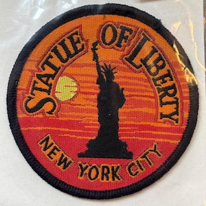 May include: Circular patch featuring the Statue of Liberty in silhouette against a sunset backdrop. The patch is embroidered with the words "STATUE OF LIBERTY" and "NEW YORK CITY" in a bold font, with a black border.