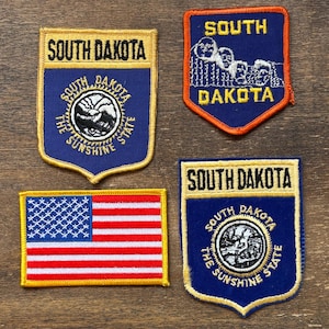 Könnte beinhalten: Vier gestickte Aufnäher mit dem Text "SOUTH DAKOTA". Ein Aufnäher zeigt die amerikanische Flagge. Zwei Aufnäher haben ein kreisförmiges Design mit Text. Ein Aufnäher zeigt Mount Rushmore. Alle Aufnäher haben einen goldenen Rand.
