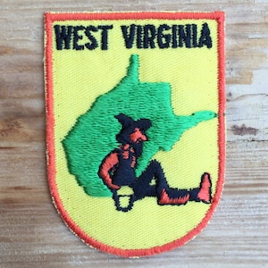 Op de afbeelding: Geborduurde patch met de tekst "WEST VIRGINIA" in zwart boven een groene omtrek van de staat. Een cartoonfiguur in zwart en oranje ligt op de groene vorm, tegen een gele achtergrond met een rode rand.