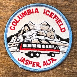 Könnte beinhalten: Ein runder Aufnäher mit hellblauem Rand. Er zeigt die Worte "COLUMBIA ICEFIELD" und "JASPER, ALTA." in Rot. Der Aufnäher zeigt eine Berglandschaft mit einem rot-weißen Bus.