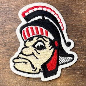 Puede incluir: Un parche bordado que presenta una cabeza de mascota estilizada con un casco de estilo romano en negro y rojo. El diseño incluye una cara de color crema con contornos negros, una cresta a rayas rojas y blancas y un borde blanco alrededor.
