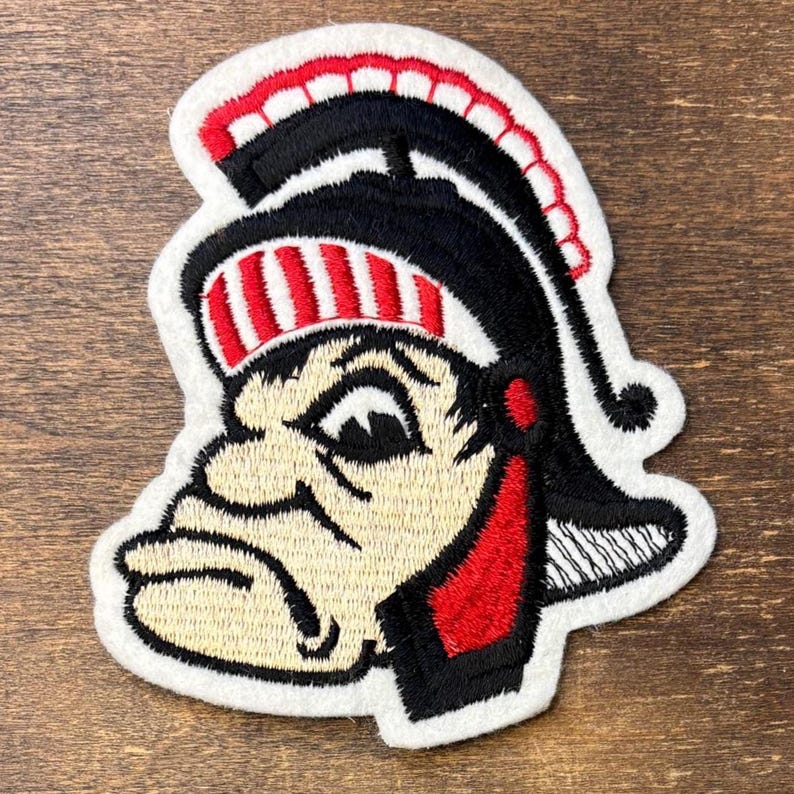 Pu&ograve; includere: Una toppa ricamata raffigurante una testa di mascotte stilizzata che indossa un elmo in stile romano nero e rosso. Il design include un volto color crema con contorni neri, una cresta a strisce rosse e bianche e un bordo bianco attorno.