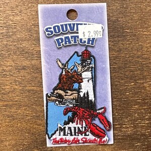 May include: A souvenir patch featuring the state of Maine. The patch includes a moose, a lighthouse, and a lobster, with the word "MAINE" and the phrase "The Way Life Should Be" embroidered on it. The patch is attached to a purple backing.