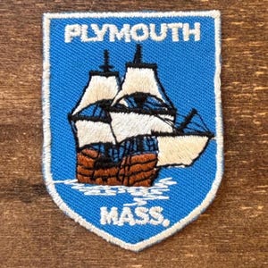 May include: A blue and white embroidered patch with the text "PLYMOUTH MASS." and an image of a tall ship. The patch has a shield shape with a white border and is likely a souvenir or collectible item.