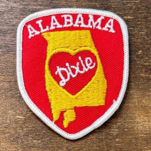 May include: A shield-shaped embroidered patch with a red background and a white border. It features the word ALABAMA in white capital letters at the top, a yellow outline of the state of Alabama, and a red heart containing the word Dixie in white.