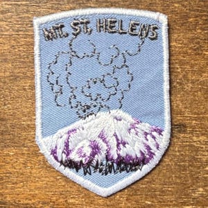 May include: Embroidered patch featuring Mt. St. Helens. The patch has a light blue background with the mountain in white and purple. The text "MT. ST. HELENS" is embroidered at the top in a dark blue color.