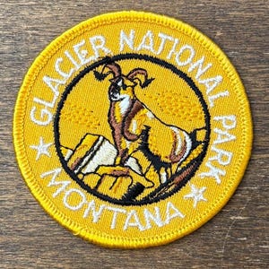 Può includere: Una toppa rotonda gialla del Glacier National Park. La toppa presenta un camoscio marrone e bianco su un paesaggio roccioso, circondato da un cerchio nero. Le parole "Glacier National Park Montana" sono ricamate in bianco.