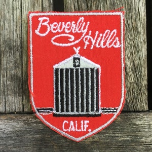 Puede incluir: Un parche bordado en forma de escudo con fondo rojo y borde blanco. El texto Beverly Hills está bordado en letra cursiva blanca en la parte superior, y CALIF. en blanco en la parte inferior. Un gráfico de parrilla de coche en blanco y negro.