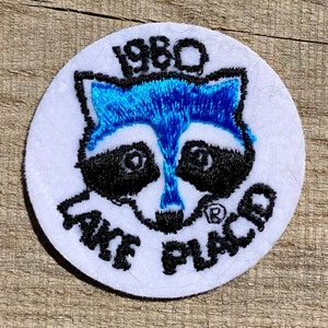 Puede incluir: Un parche circular blanco con texto bordado en negro que dice "1980 Lake Placid" y una cara de mapache bordada en negro y azul.
