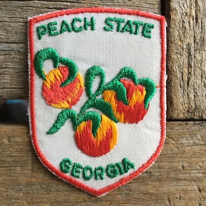 May include: Embroidered patch featuring three peaches with red and yellow fruit and green leaves. The patch reads "PEACH STATE GEORGIA" in green lettering, outlined in red, on a white background.