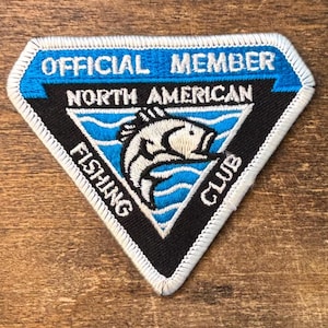 Puede incluir: Un parche bordado triangular con borde blanco. La sección azul superior dice OFFICIAL MEMBER. El centro negro presenta un gráfico de un pez blanco sobre olas azules, con el texto NORTH AMERICAN FISHING CLUB alrededor del diseño.