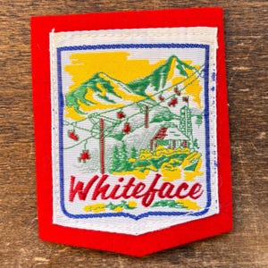 May include: A rectangular patch with a red border and a white interior. The patch features a mountain scene with a ski lift, trees, and a building. The word "Whiteface" is written in red script at the bottom.