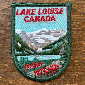 May include: Embroidered patch featuring a scenic view of Lake Louise, Canada. The patch has a light blue background with a mountain range, lake, and red flowers. The text "LAKE LOUISE CANADA" is in red.