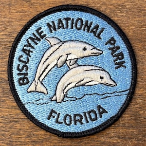 May include: Embroidered patch featuring two leaping dolphins in a light blue ocean scene. The patch is circular with a black border and text that reads "BISCAYNE NATIONAL PARK FLORIDA" in black lettering.