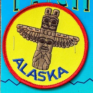 May include: Embroidered patch featuring a brown totem pole with outstretched wings, set against a yellow background. The patch has a red border and the word "ALASKA" in blue lettering. The design is circular.