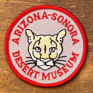 May include: Round patch with a red border and the text "ARIZONA-SONORA DESERT MUSEUM". The patch features a cougar's face in yellow and white on a gray background.