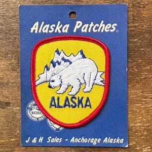 Puede incluir: Parche de Alaska amarillo y rojo con un oso polar y montañas. La palabra "ALASKA" está en azul. El parche está sobre una tarjeta azul con las palabras "Alaska Patches" en la parte superior.
