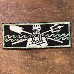 May include: A rectangular embroidered patch featuring a black background with a central white figure of a bearded king wearing a crown. Behind the figure are a white shovel and a white trident crossed, set against stylized light green ocean waves.