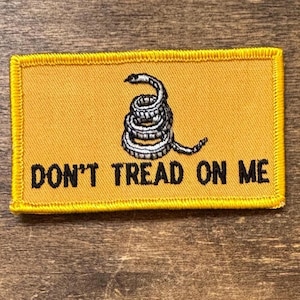 May include: A rectangular yellow patch with a coiled rattlesnake in black and white. The text "DON'T TREAD ON ME" is printed in black, below the snake. The patch has a yellow border.
